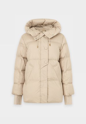 Une femme porte une doudoune beige à capuche sur un pull à motif argyle, un pantalon crème et des chaussures à talons pointus, sur un fond clair uni.