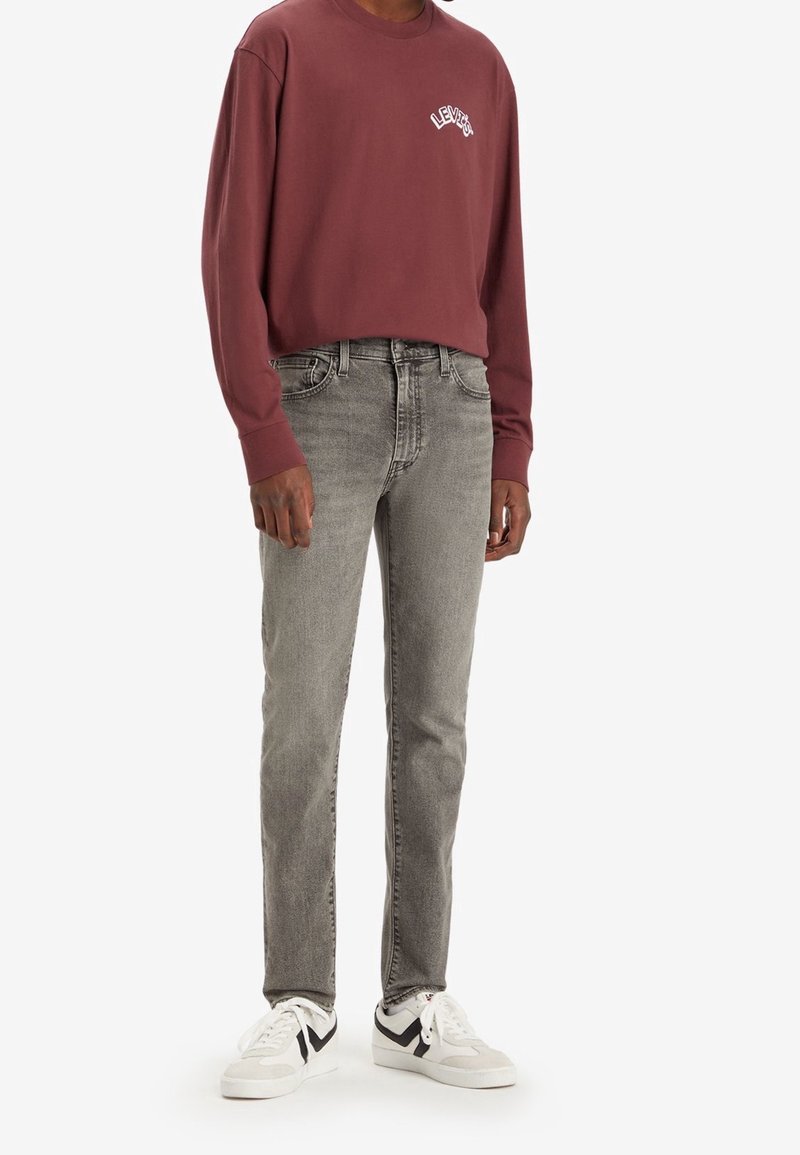 Personne portant un t-shirt à manches longues Levi's bordeaux, un jean slim gris et des baskets blanches avec des accents noirs, debout devant un fond blanc.