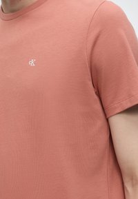 Κοντινό πλάνο ενός ατόμου που φοράει ένα απαλορόζ κοντομάνικο t-shirt με μικρό λευκό λογότυπο "cK" κεντημένο στο στήθος.