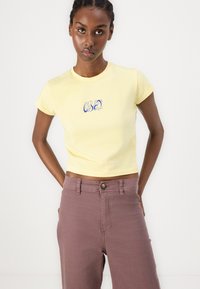Kollane lühike T-särk sinise "OBEY" logoga, kombineeritud kõrge vöökohaga tuharoosade pükstega. Lühikesed varrukad, pehme puuvillane materjal.
