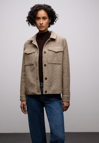 Béžová texturovaná bunda se dvěma náprsními kapsami a zapínáním na knoflíky, v kombinaci s tmavým topem a modrými širokými džíny.