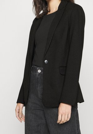 Personne portant un blazer noir sur un haut noir et un jean en denim gris foncé, se tenant devant un fond clair uni.