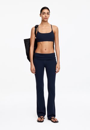 Vrouw draagt een marineblauwe sportbeha, bijpassende yogabroek, teenslippers en draagt een zwarte schoudertas, staand tegen een witte achtergrond.