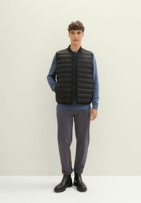 Schwarze gepolsterte Weste mit Stehkragen, die über einem blauen Langarmshirt getragen wird. Das Modell kombiniert sie mit grauen hochgekrempelten Hosen und schwarzen Stiefeln.