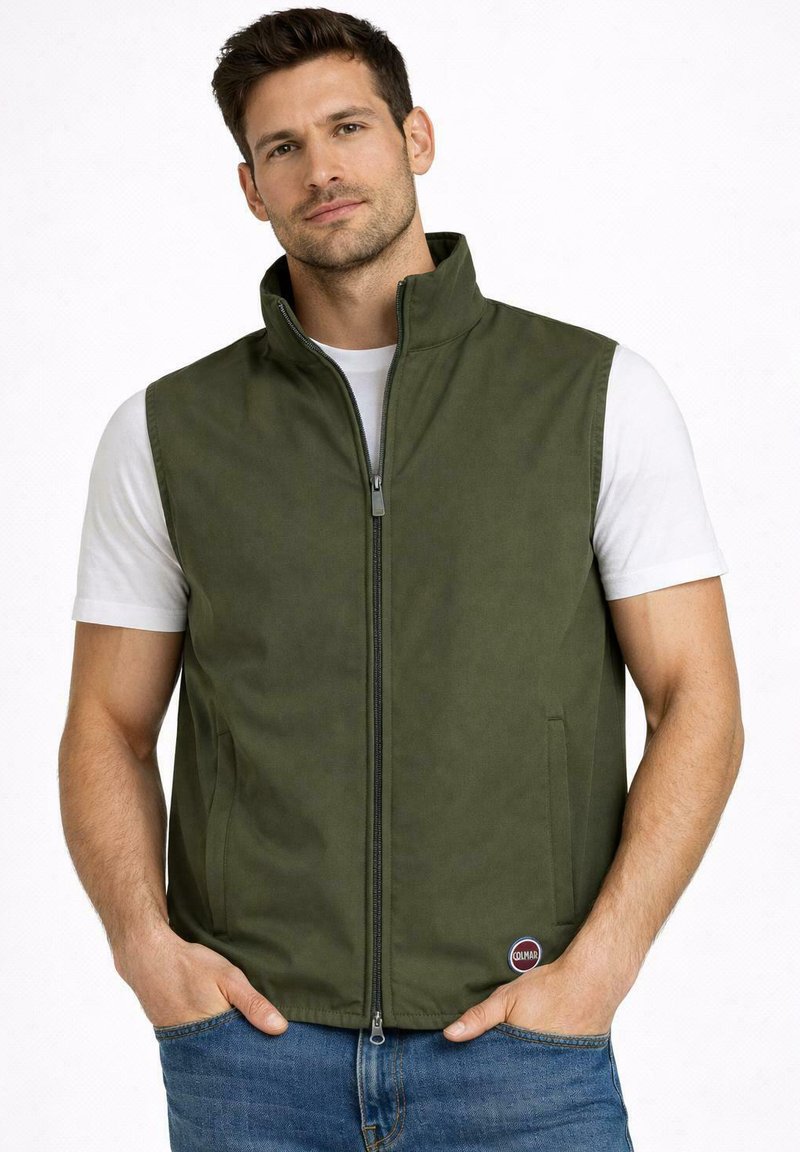 Uomo con capelli corti e scuri che indossa un gilet senza maniche verde oliva con cerniera sopra una t-shirt bianca e jeans blu, con le mani nelle tasche del gilet.