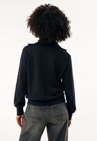 Sudadera negra con cuello, con mangas largas y un corte holgado. Combinada con jeans grises que muestran bolsillos traseros.