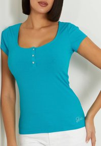 Donna che indossa una maglietta aderente di colore turchese a maniche corte e costine, con tre bottoni decorativi vicino allo scollo, e pantaloni bianchi.