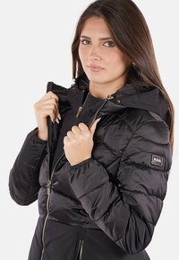 Giacca a vento nera con cappuccio, caratterizzata da sezioni trapuntate, chiusura con cerniera e polsini elasticizzati. Piccola etichetta con logo sul lato sinistro.