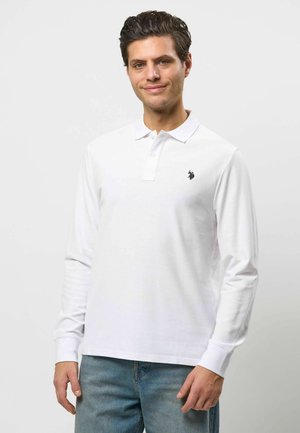 Muž s tmavými vlasmi, oblečený v bielom polo tričku s dlhým rukávom a malým čiernym logom na hrudi, modré džínsy, mierne sa usmieva.