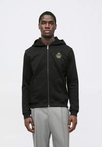 Capa preta com fecho e capuz, feita de tecido macio, com bolso frontal. Destaque para o logótipo dourado no peito.