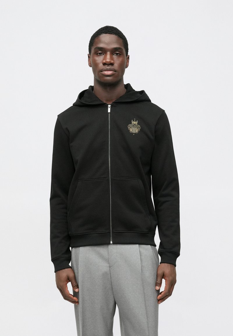 Capa preta com fecho e capuz, feita de tecido macio, com bolso frontal. Destaque para o logótipo dourado no peito.