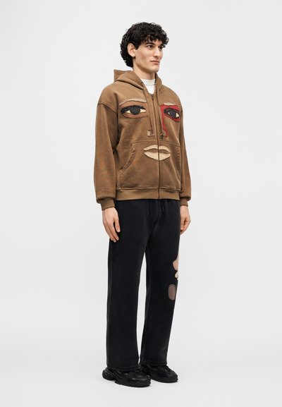 Καφέ φούτερ με κουκούλα και σχεδιασμό προσώπου· αντίθετα κεντημένα μάτια και στόμα. Συνδυασμένο με μαύρο φόρμας παντελόνι με patches και στενή εφαρμογή.