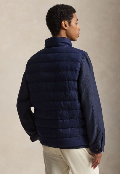 Mężczyzna w granatowej pikowanej kamizelce na niebieskiej koszuli z długim rękawem i beżowych spodniach, stojący bokiem na neutralnym tle.