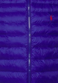 Fialová nafukovací vesta s horizontálním švem, předním zipem a malým červeným logem. Hladká, lesklá tkanina s lehkým a izolovaným designem.