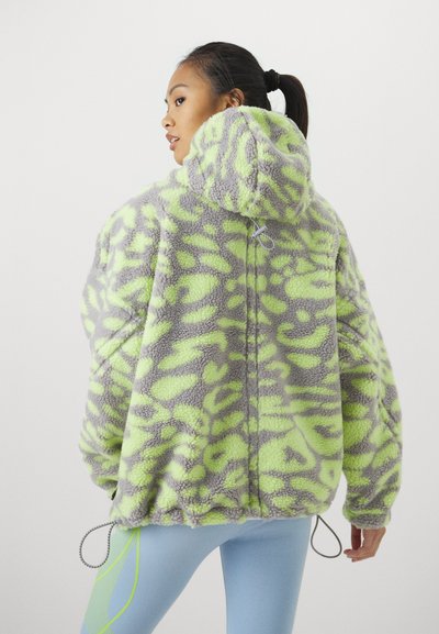 Mulher com cabelo apanhado a usar uma jaqueta de fleece cinza e verde néon com capuz, combinada com leggings azul claro com detalhes em verde néon.