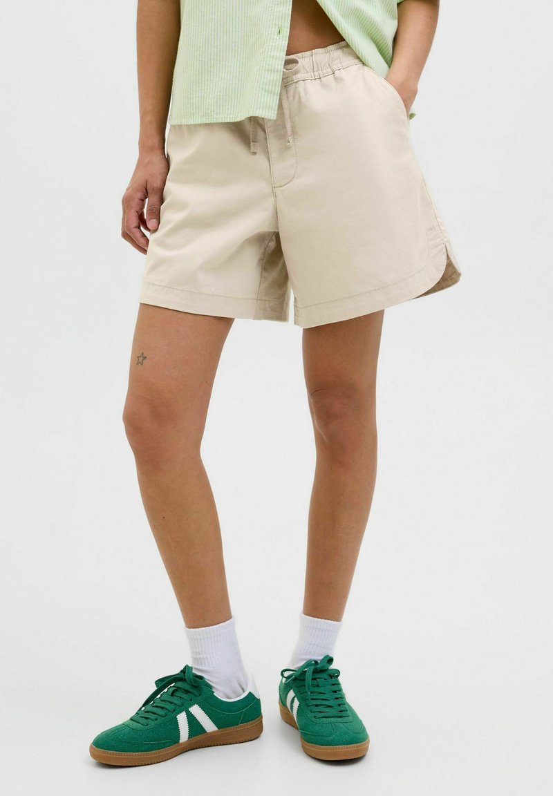 Pantalones cortos de algodón beige con cintura elástica y bolsillos laterales, con un ligero curvado en el dobladillo. Combinados con zapatillas verdes y calcetines blancos.
