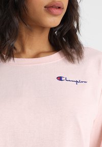 Kvinde med mellemlangt mørkt hår iført en lys pink Champion sweatshirt med logosyning på brystet.