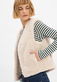 Fleece vest in lichtbeige met een zachte, gestructureerde oppervlakte. Het heeft open zijkanten en twee zijzakken, gedragen over een gestreepte lange mouw shirt.