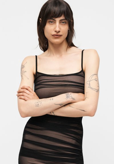 Άτομο με κοντά σκούρα μαλλιά και ορατά τατουάζ φορεί μαύρο διάφανο φόρεμα με λεπτές τιράντες, σταυρωμένα χέρια, κοιτάζει με αυτοπεποίθηση μπροστά.