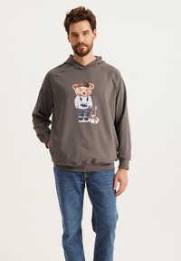 Szara bluza z kapturem z grafiką misia z aparatem, noszącego białą koszulkę i niebieskie dżinsy. W zestawie z niebieskimi jeansami, w luźnym kroju.