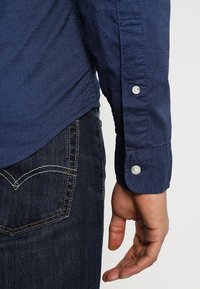 Högra armen i en blå långärmad skjorta med knäppta manschetter bredvid en mörkblå denimjeansficka med synlig vit sömnad.