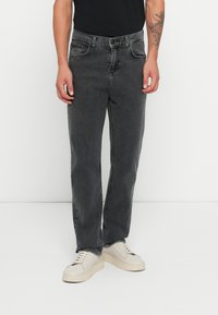 Vaqueros de mezclilla negra con corte recto y detalles de costura visibles. Combinados con zapatillas beige, de pie frente a un fondo blanco.