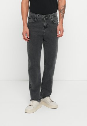 Vaqueros de mezclilla negra con corte recto y detalles de costura visibles. Combinados con zapatillas beige, de pie frente a un fondo blanco.