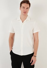 Kortærmet skjorte i off-white med vertikal ribbet tekstur, spids krave og foran knaplukning. Sæt sammen med sorte bukser.