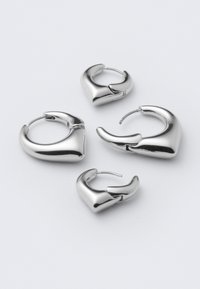 Quatre boucles d'oreilles en argent poli en forme d'anneaux épais, incurvés et légèrement angulaires, disposées sur une surface blanche.