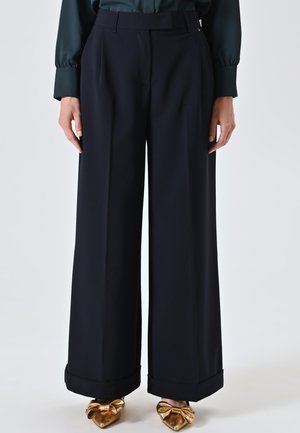 Pantaloni ampi di colore blu navy con una vestibilità sartoriale, orli cuffi e tasche laterali, abbinati a scarpe lucide con dettaglio a fiocco dorato.
