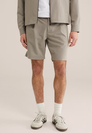 Férfi bézs rövidnadrágban, fehér zokniban, fekete csíkos fehér sportcipőben, bézs dzsekiben fehér inggel alatta.