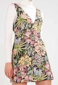 Robe sans manches en tapisserie florale avec de grandes fleurs roses, jaunes et vertes portée sur un haut col roulé blanc.