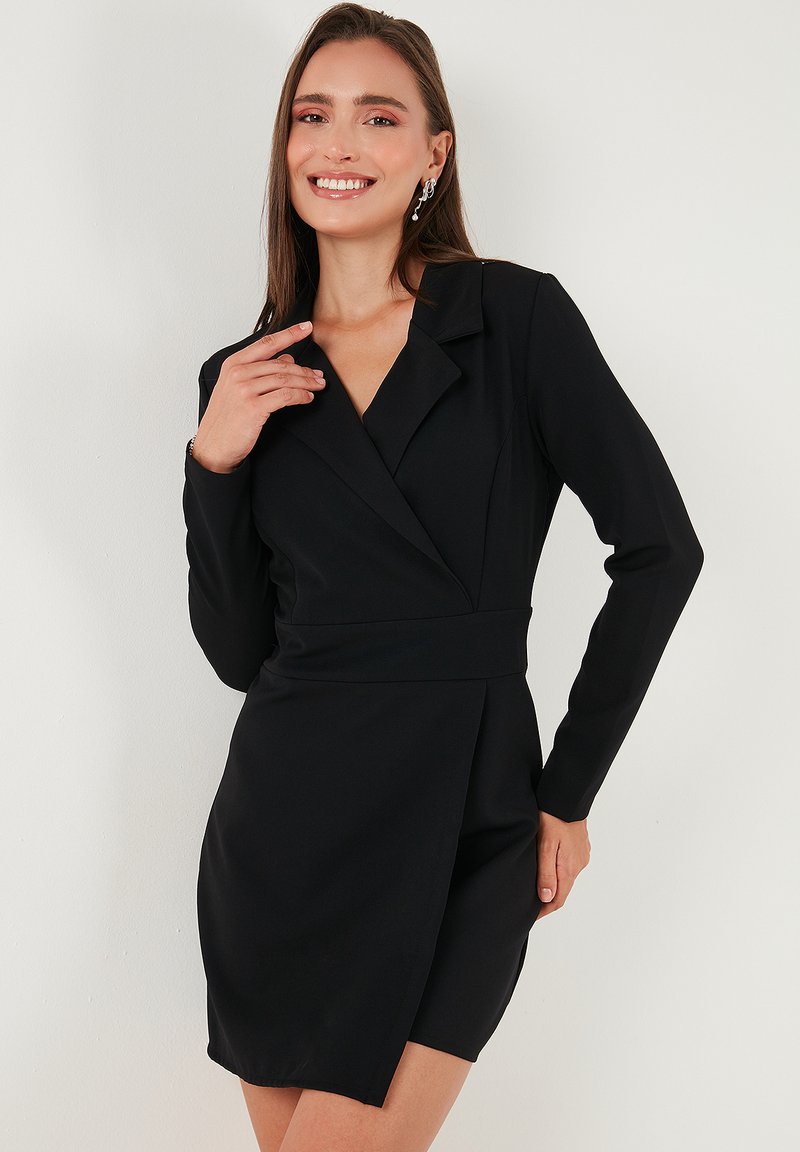 Frau mit einem Lächeln, die ein schwarzes langärmliges Blazer-Kleid mit asymmetrischem Saum und silbernen hängenden Ohrringen trägt, steht vor einer weißen Wand.