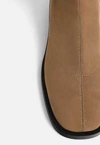 Stivale alla caviglia in suede marrone con texture liscia, punta rotonda e suola in gomma nera. Presenta una cucitura centrale e dettagli di cucitura minimalisti.