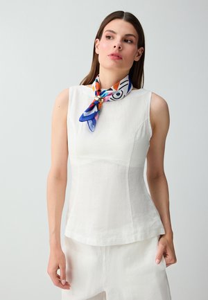 Donna con top bianco senza maniche e pantaloni, con una sciarpa colorata e decorata legata intorno al collo, su uno sfondo uniforme.