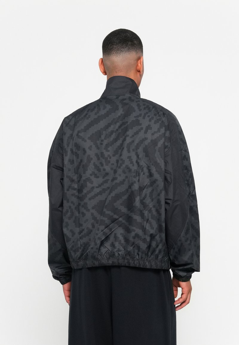 Chaqueta cortavientos negra con un patrón texturizado, puños y dobladillo elásticos, cuello alto, que muestra un ajuste relajado y una tela ligera.