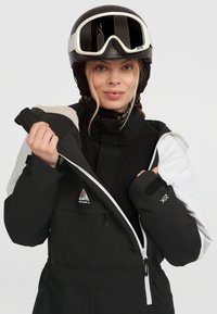 Chaqueta de nieve en blanco y negro con un cuello alto, bolsillo con cremallera y logo. Presenta tejido texturizado y puños ajustables con velcro.
