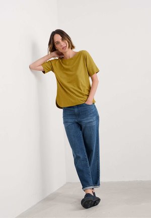 Short-sleeve, mustard-colored top with a relaxed fit, paired with light blue, loose-fitting jeans and black sneakers. Neutral background.