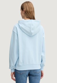 Světle modrá mikina s kapucí, volného střihu, s žebrovanými manžetami a lemem. Měkký materiál, minimální detaily šití a bez viditelného loga.