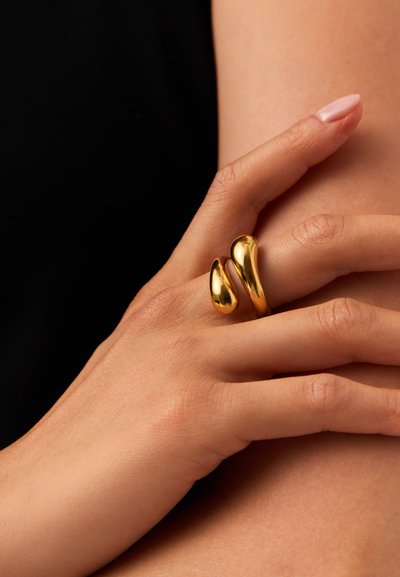 Χέρι με απλό χρυσό ανοιχτό δαχτυλίδι που έχει δύο στρογγυλεμένες, σχήματος σταγόνας άκρες, που ακουμπούν πάνω σε γυμνό δέρμα με μαύρο φόντο.