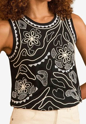 Czarny top bez rękawów z białymi haftowanymi kwiatowymi i abstrakcyjnymi wzorami, noszony z jasnymi spodniami.