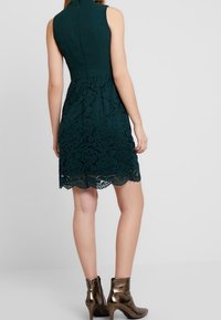 Ärmelloses dunkelgrünes Kleid mit tailliertem Oberteil und einem Spitzenrock mit floralen Mustern. Mit gewelltem Saum und Reißverschluss hinten.