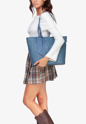 Borsa a tote in pelle sintetica blu con un accento a nastro blu trapuntato, forma rettangolare e chiusura con zip, indossata da un modello con una gonna a quadri.