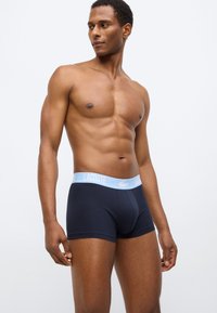 Boxers pour hommes bleu marine avec une ceinture élastique arborant un logo Lacoste bleu clair. Fabriqués en tissu élastique, offrant une texture lisse et un ajustement confortable.