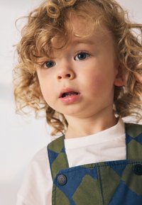 Enfant portant une chemise blanche sous une salopette à motifs verts et bleu marine. Cheveux bouclés et fond neutre mettant en valeur les couleurs et le design de la tenue.