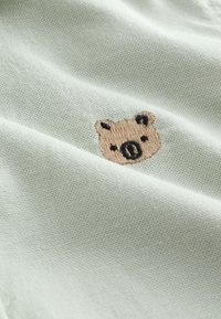 Light gray fabric with small embroidered brown bear face with black details, visible texture and folds in the cloth.