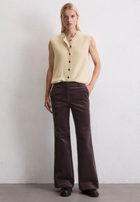 Crème mouwloze top van gestructureerde stof met een knoopsluiting, gecombineerd met donkerbruine flared corduroy broeken en zwarte schoenen.