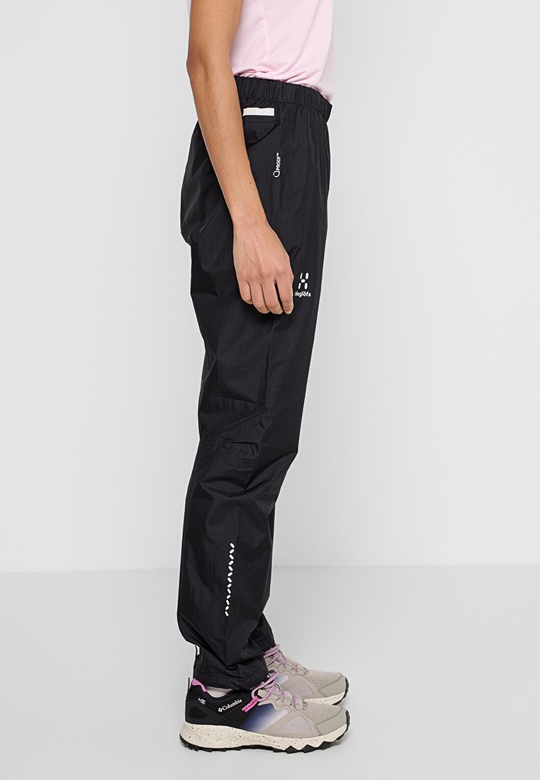 Mustat vedenpitävät housut, joissa on joustava vyötärö, sivutaskut ja heijastavat yksityiskohdat; yhdistetty vaaleanharmaisiin ja vaaleanpunaisiin vaelluskengiin.