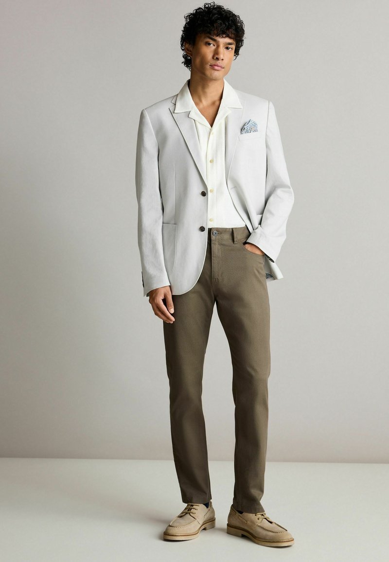 Lys grå blazer over en hvid skjorte med krave, parret med brune bukser og beige sko. Lommenæseklud tilføjer et mønstret accent.