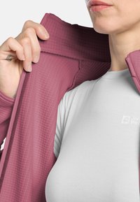 Veste en polaire de couleur rose pastel avec une surface texturée, associée à une chemise ajustée gris clair avec un logo sur la poitrine.